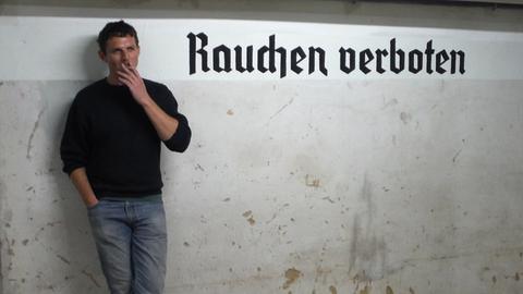 Martin Wenzel steht mit einer Zigarette in der Hand vor einem Schriftzug an der Wand "Rauchen verboten"