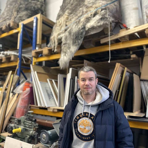Felix Große-Lohmann, Gründer von "Material für alle", steht in einem Lagerraum. Hinter ihm sind Holzbalken, Felle, Bilderrahmen und andere Materialien in Regalen zu sehen.