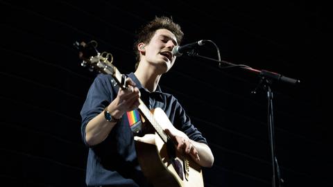 Un giovane in piedi sul palco con la sua chitarra in spalla.