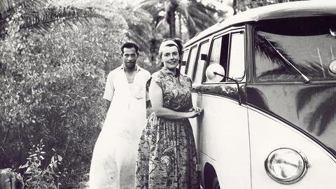 Eine Frau steht neben einem alten VW-Bus, im Hintergrund ein Mann mit weißem Gewand