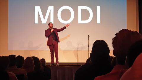Mann mit Mikrofon auf der Bühne, im Hintergrund der Schriftzug MODI.