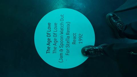 Das Bild zeigt einen Blick auf den Boden im Frankfurter Techno-Museum MOMEM: Darauf ist ein türkisfarbener Kreis angebracht mit den Eckdaten zum Song "The Age of Love" von 1992. Zwei in schwarze Stiefel gekleidete Füße stehen davor.