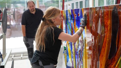 Ein Frau arbeitet mit Handschuhen an einem bunten Glas-Kunstwerk. Im Hintergrund schaut ein Mann zu. 