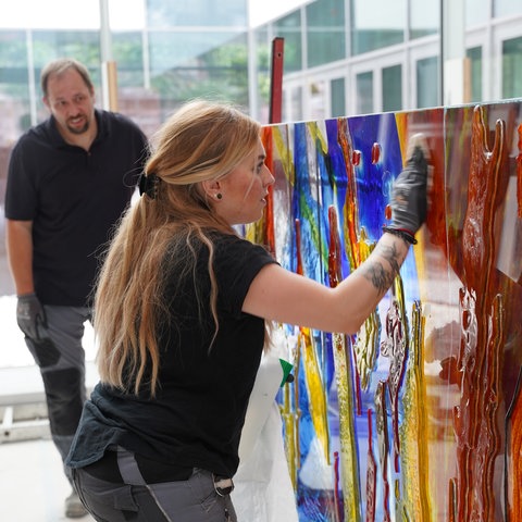 Ein Frau arbeitet mit Handschuhen an einem bunten Glas-Kunstwerk. Im Hintergrund schaut ein Mann zu. 