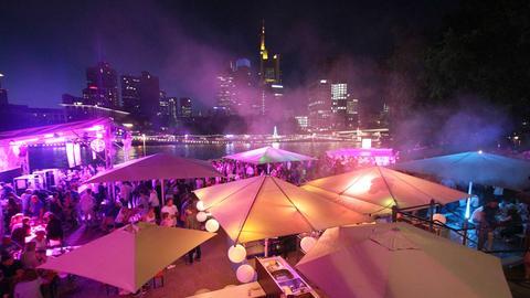 Bunt beleuchtete gastronomische Stände im Bildvordergrund und die Skyline Frankfurts im Hintergrund.