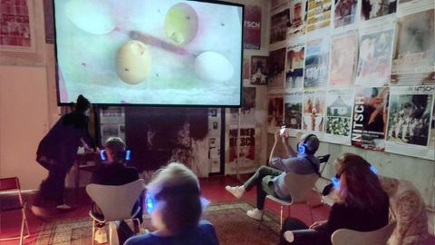 In einem abgedunkelten Raum sitzen Menschen mit OP-Masken und blau leuchtenden Kopfhörern auf Stühlen und schauen auf eine temporäre Leinwand, die mitten in einem kleinen Raum mit vielen Plakaten an der Wand steht und ein Musikvideo zeigt. Auf der Leinwand sind Eier zu sehen.