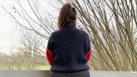 Das Bild zeigt eine Frau von hinten, die auf einem Balkon steht. Sie trägt einen dunklen Zopf und blauen Strickpulli.