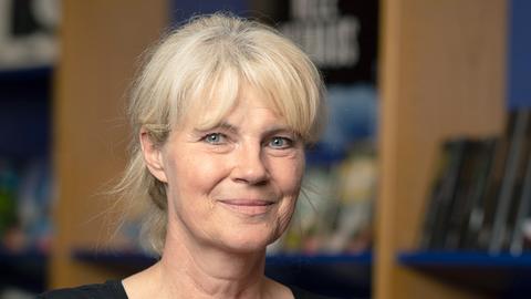 Eine Frau mit blonden Haaren und schwarzem T-Shirt steht vor einem Bücherregal.