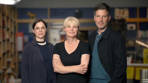 Zwei Frauen und ein Mann stehen mit verschränkten Armen nebeneinander und blicken in die Kamera.