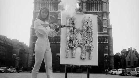 Das Bild zeigt Künstlerin Niki de Saint Phalle in einem weißen Ganzkörperanzug und mit schwarzen Stiefeletten vor Notre-DAme in Paris. Sie hält ein Gewehr in der Hand. Neben ihr ist ein Bild auf einer Staffelei zu sehen.