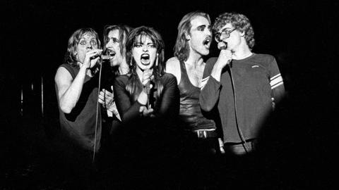 Das Schwarz-Weiß-Bild zeigt die Nina Hagen Band bei einem Auftritt in der Stadthalle Braunschweig im Jahr 1979. In der Mitte ist Sängerin Nina Hagen zu sehen, sie hat den Mund offen und ein Mikrofon in der Hand. Links und rechts von ihr stehen jeweils zwei männliche Bandmitglieder und singen gemeinsam in jeweils ein Mikrofon.