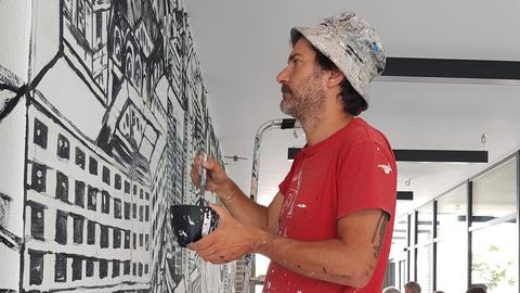 Ein Mann in rotem T-Shirt und mit Hut steht auf einer Leiter und malt ein schwarz-weißes Bild von Offenbach an eine Garagenwand.