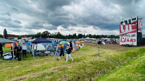 Drei junge Männer laufen über den Festival-Campingplatz. Im Hintergruund sind Zelte zu sehen, rechts ein Turm mit einem Open Flair Plakat zu sehen.