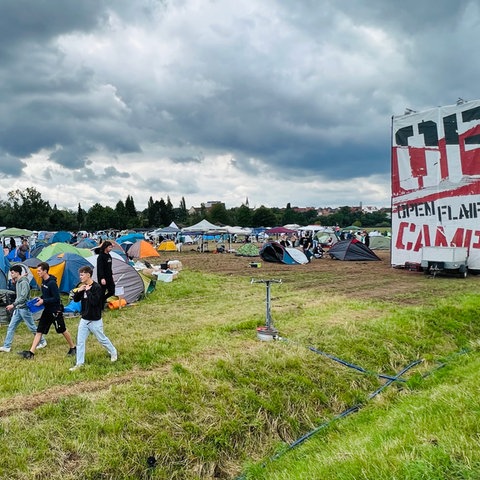 Drei junge Männer laufen über den Festival-Campingplatz. Im Hintergruund sind Zelte zu sehen, rechts ein Turm mit einem Open Flair Plakat zu sehen.