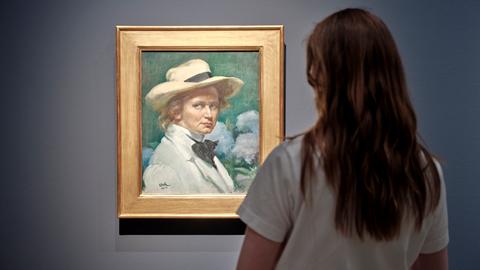 Das Bild zeigt die Rückansicht einer Frau mit weißem T-Shirt und langen, dunklen Haaren, die vor einem Selbstporträt der Malerin Ottilie W. Roederstein steht.