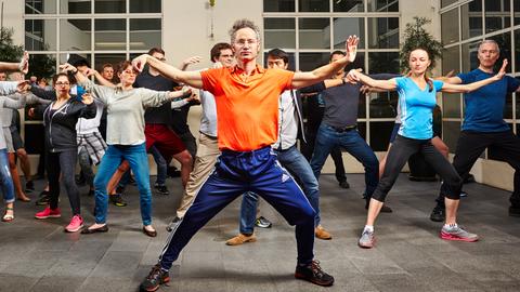 Ein Mann im orangenen Tshirt steht vor einer Gruppe und macht Tai Chi-Bewegungen.