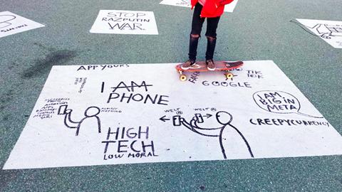 Eine Skateboarderin fährt über Kunstwerke am Boden: schwarze geschriebene Texte auf weißen Rechtecken, z.B. "High tech, low moral" oder "I AM PHONE"