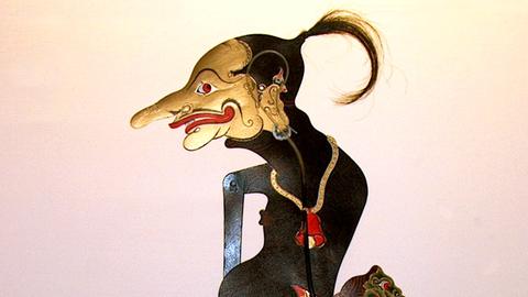 Das Bild zeigt eine Puppe aus einem Schattentheater. Sie hat ein goldenes Gesicht mit einer langen Nase und wülstigen Lippen, einen dunklen Pferdeschwanz und einen schwarzen dicken Bauch.