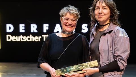Zwei Frauen halten einen goldenen Barren mit der Aufschrift "DERFAUST" in den Händen
