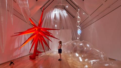 Das Bild zeigt eine Ausstellungsansicht von "Plastic World" in der Schirn. Eine Frau steht vor einem großen roten Stern aus Kunststoff, hinter ihr sind transparente Bälle.