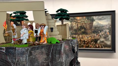 Playmobilfiguren in Kleidung der Antike stehen auf einem Plastikfelsen vor einem Gemälde