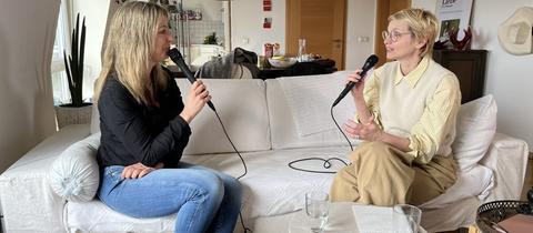 Zwei Frauen sitzen auf einem Sofa, ein Mikrofon in der Hand