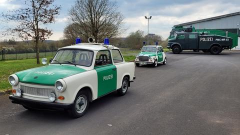Das Bild zeigt einen grün-weiß gemusterten Trabbi mit der Aufschrift "Polizei" an den Seiten. Im Hintergrund sind weitere Polizei-Oldtimer zu sehen, darunter ein grün-weißer Fiat 500 und ein schwarzer Wasserwerfer.