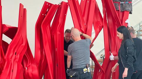 Das Bild zeigt eine Skultpur der Künstlerin Bettina Pousttchi. Sie besteht aus roten Leitplanken, die verschieden gebogen und längs aufgestellt sind. Drei Männer und eine Frau in dunkler Kleidung installieren sie im Museum Reinhard Ernst in Wiesbaden.