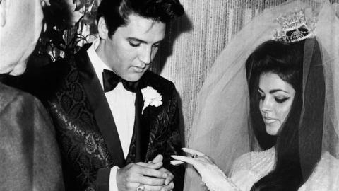 Das Bild zeigt Elvis und Priscilla Presley bei ihrer Hochzeit. Er trägt einen schwarzen Anzug und schwarze Haare und schaut nach unten, um ihr den Ring an den Ringfinger zu stecken. Sie hat dunkle, auftoupierte Haare und trägt einen Schleier. 