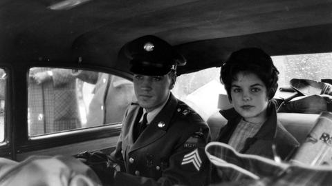 Das Bild zeigt Elvis und Priscilla Presley 1960 in Bad Nauheim. Sie sitzen in einem Taxi. Er trägt eine Militäruniform, Priscillas Haare sind auftoupiert. Sie trägt einen Mantel uind ein kariertes Kleid.