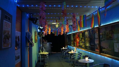 Ein dunkler Raum mit mehreren Sitzgruppen aus blauen Bistrostühlen und -tischen. An den Wänden hängen Filmplakate, an der Decke Blumenketten und eine Girlande in Regenbogenfarben.
