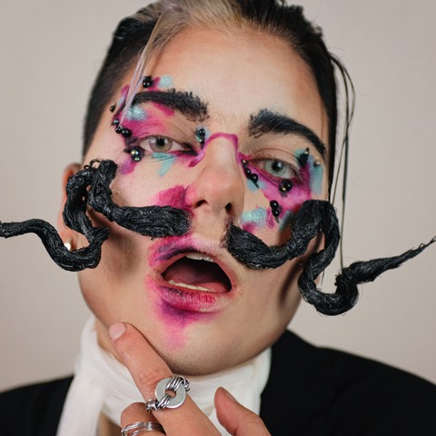 Das Bild zeigt eine Person, die direkt in die Kamera blickt. Der Mund ist geöffnet und pink verschmiert geschminkt. Die weißblond und schwarz gefärbten Haare sind zurückgegelt, die Augen lila und türkis geschminkt. Die Person trägt außerdem einen auffällig nach oben gezwirbelten schwarzen Schnurrbart. 