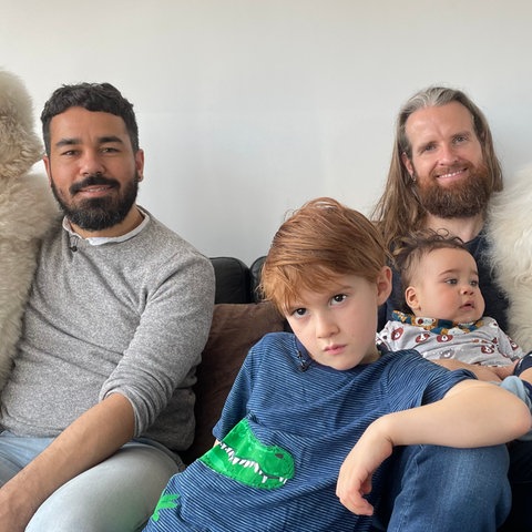 Zwei bärtige Männer sitzen mit zwei jüngeren Kindern und zwei Pudeln auf dem Sofa.