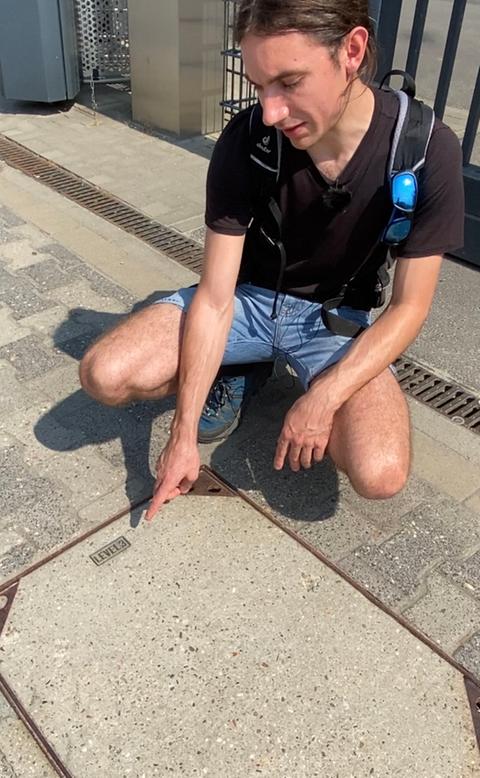 Ein Mann zeigt hockernd auf eine im Boden eingelassene Betonplatte
