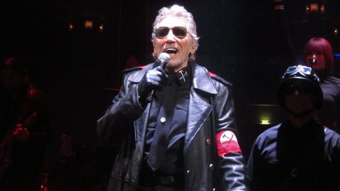Roger Waters in einem schwarzen Outfit mit Armbinde mit zwei gekreuzten Hämmern darauf.