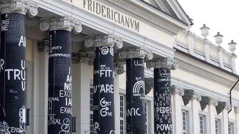 Mattschwarz gestrichene Säulen vor einem historischen Gebäude sind mit weißen Buchstaben, Strichmännchen und Symbolen gestaltet.