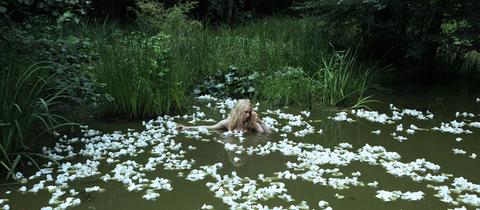 Eine Frau badet in einem Teich, scheinbar umgeben von Seerosen, die aber aus zerdrückten Plastikbechern bestehen.