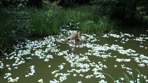 Eine Frau badet in einem Teich, scheinbar umgeben von Seerosen, die aber aus zerdrückten Plastikbechern bestehen.