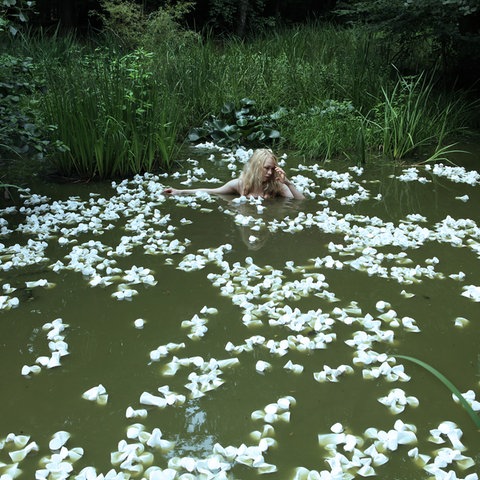 Eine Frau badet in einem Teich, scheinbar umgeben von Seerosen, die aber aus zerdrückten Plastikbechern bestehen.