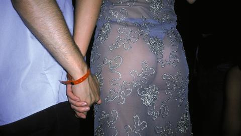 Ein händchenhaltendes Paar von hinten, die Frau trägt ein durchsichtiges Kleid.