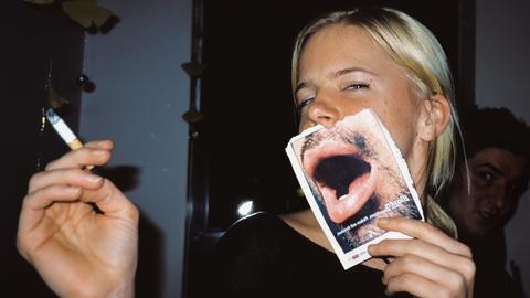 Eine junge Frau mit einer Zigarette in der Hand hält sich ein Blatt vor den Mund, auf dem ein halbes Männergesicht zu sehen ist.