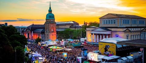 Menschenmasse zwischen Gebäuden, Zelten und Bühne in der Innenstadt Darmstadts - aus der Froschperspektive fotografiert. Im Hintergrund ein leuchtender Abendhimmel.