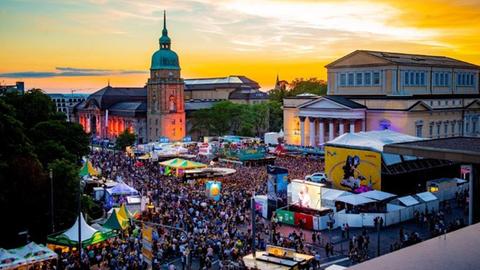 Menschenmasse zwischen Gebäuden, Zelten und Bühne in der Innenstadt Darmstadts - aus der Froschperspektive fotografiert. Im Hintergrund ein leuchtender Abendhimmel.