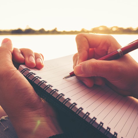 Ein Person sitzt an einem See und schreibt in ein liniertes Ringbuch.