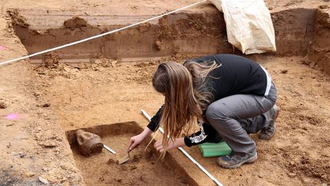 Das Bild zeigt eine archäologische Grabungsstätte. Zu sehen ist eine Frau mit langen braunen Haaren, die in einem ausgehobenen, von Lehm umgebenen Bereich hockt. Sie hält einee Art Metallspatel in der Hand, mit dem sie etwas aus dem Boden hebt.