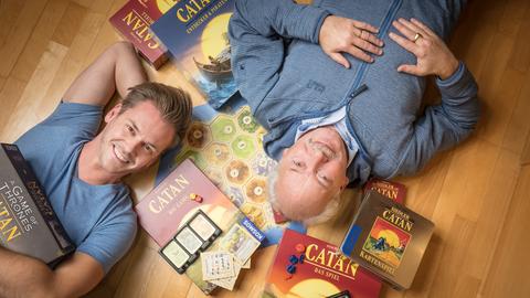 Spiele-Erfinder Klaus Teuber und sein Sohn Benjamin liegen auf dem Boden, um sie herum sind Catan-Spiele verteilt.