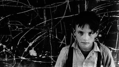 Carlos Siquier, aus der Serie "La Chanca", Almería, 1960, Schwarz-Weiß-Aufnahme, ein Junge mit schwarzen Haaren blickt von unten in die Kamera, er trägt ein Hemd und Hosenträger