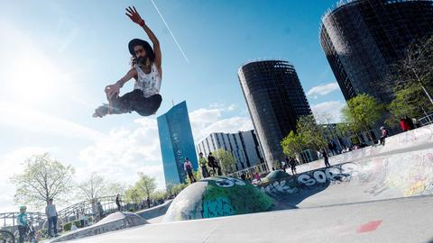 Ein Skater fliegt mit seinem Brett durch die Luft. Im Hintergrund ein Park und etwas Skyline von Frankfurt.