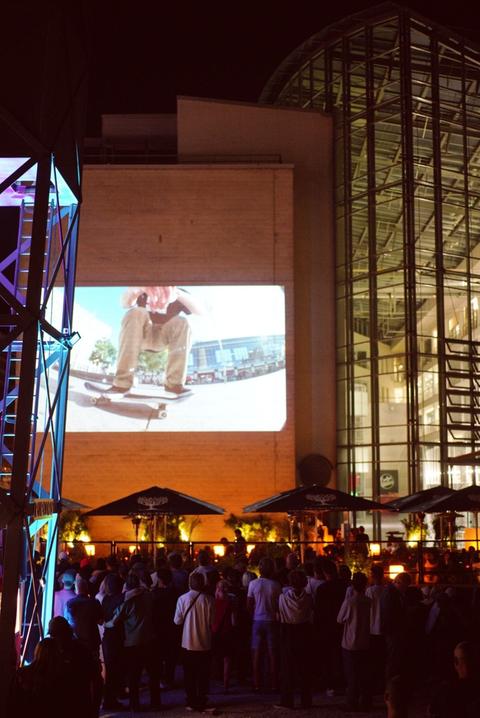 Das Bild zeigt ein Skate-Video auf einer Leinwand im Freien, davor Publikum.