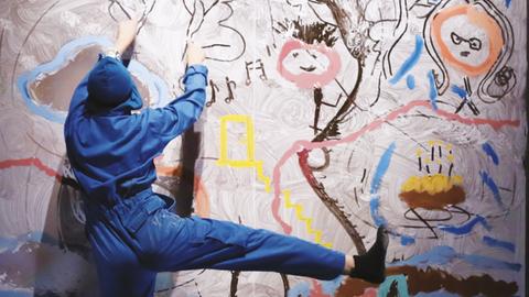 Mensch im Blaumann steht auf einer Leiter vor einer bunten Wand